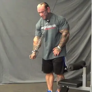 A muscular man in a gym performs a tricep workout using cable pulleys. He wears a gray t-shirt, black shorts, and blue sneakers. The background features a gray curtain and gym equipment.