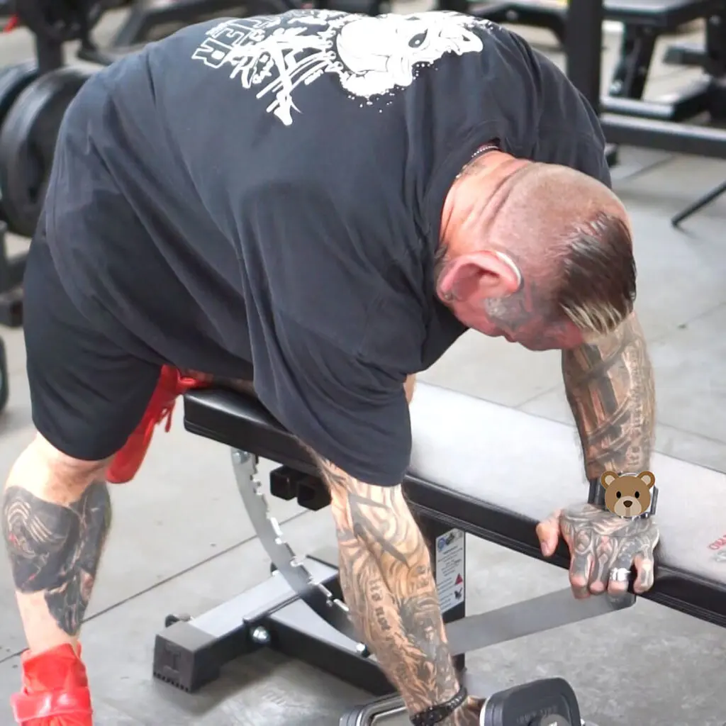 A heavily tattooed man leans over a gym bench, gripping a dumbbell with one hand. He is wearing a black t-shirt, red shorts, and red shoes. Gym equipment is visible in the background.