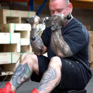 A heavily tattooed man is seated and performing a bicep workout using a cable machine in a gym. He is wearing a black T-shirt, black shorts, and red shoes. Wooden crates are visible in the background.
