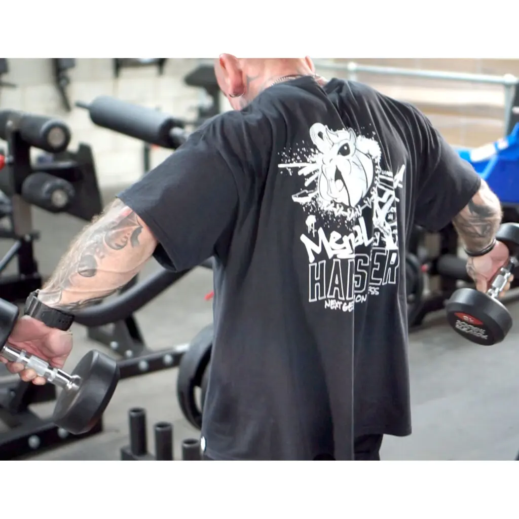 A person with tattoos is lifting dumbbells in a gym. They are wearing a black t-shirt with a graphic design on the back. Gym equipment is visible in the background.