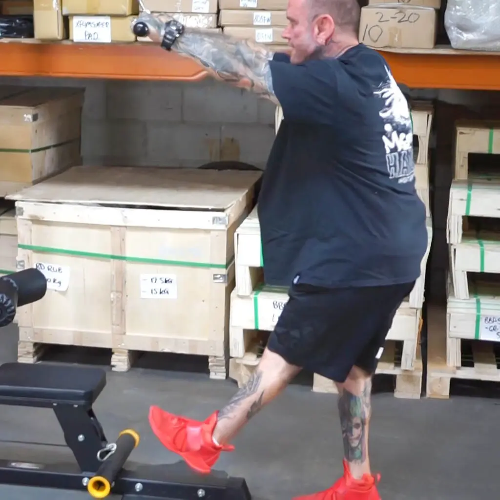 A person in a gym performs a cable pull exercise with one leg raised, wearing bright red shoes and a black shirt adorned with white text. Wooden crates are stacked on shelves in the background.