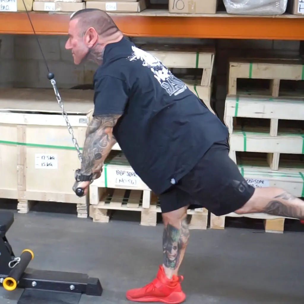 A heavily tattooed person in a black shirt exercises in a warehouse, performing a strength training move with a cable machine. They stand on one leg, balancing with a focused posture. Wooden pallets and boxes are visible in the background.