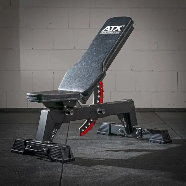 An ATX® Utility Bulls Bench, adjustable and black with a red adjustment bar, sits partially inclined on a gym floor against a white brick wall.