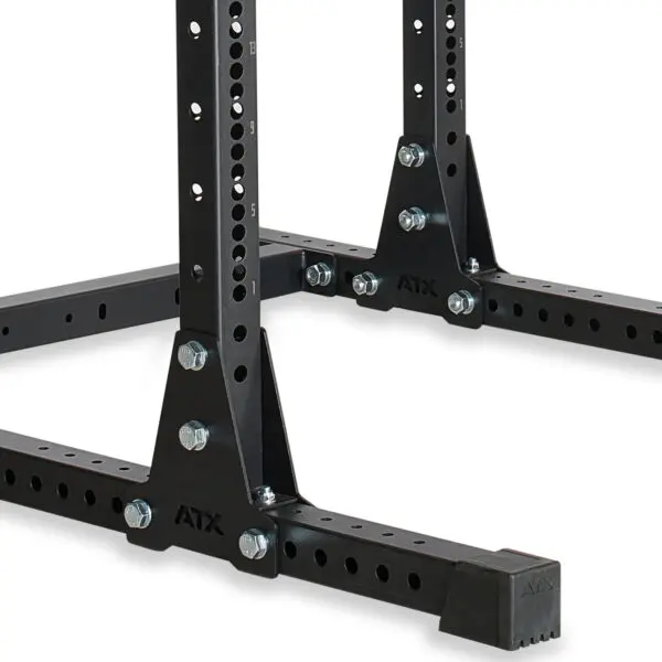 A close-up of the ATX® Power Rack 755 Short Distance shows a black metal stand with multiple holes and bolts. It features a sturdy base with a rectangular foot and rubber caps for stability, designed to support heavy equipment.
