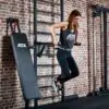 A person in workout attire performs tricep dips on parallel bars attached to the ATX® Wall Bar Gym, a wall-mounted exercise station with Swedish Bars, set in a gym with brick walls. Nearby is a padded incline bench.
