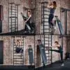 A woman in athletic wear uses the ATX® Wall Bar Gym against a brick wall to perform pull-ups, use an inclined bench, hang from stall bars, and stretch her leg.