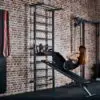 A person performs incline sit-ups on an ATX® Wall Bar Gym bench in a gym, located next to Swedish bars and other exercise equipment. Theyre wearing a black outfit and have their feet secured under a bar against the brick wall.