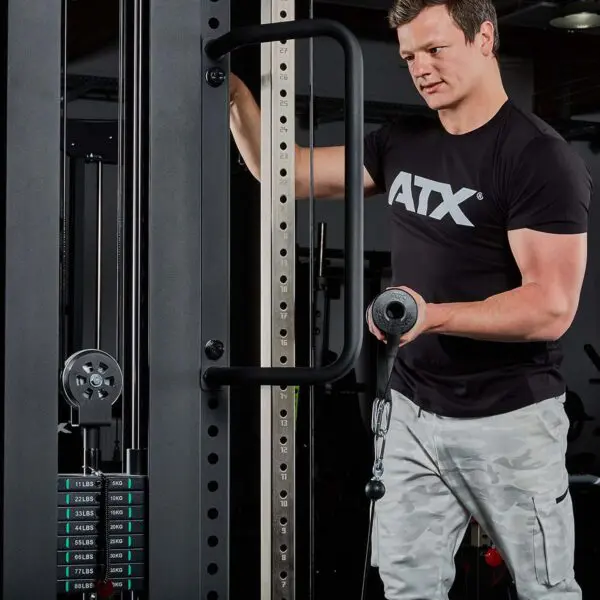 A man in an ATX tee adjusts a gym cable machine, holding a bar enhanced with ATX® Conic Fat Grips. The machine shows weight options starting at 11 lbs as he focuses on his setup.