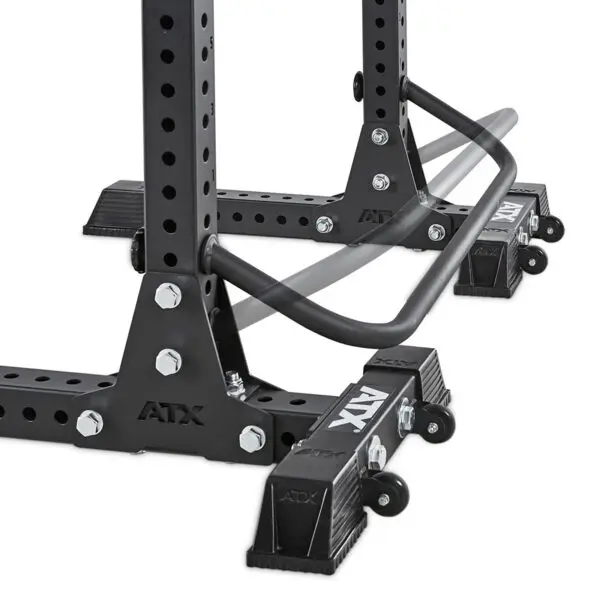 Close-up of a black ATX® Squat Stands Brace with adjustable safety arms. The rack features customizable holes and large bolts for stability, with foot caps securing the base. A partially blurred safety bar shows movement.