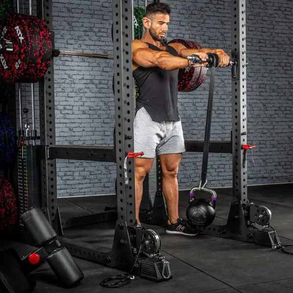 A man in a gym exercises with the ATX® Forearm Training Set, utilizing resistance bands fixed to a squat rack. Hes dressed in a black tank top and gray shorts, surrounded by diverse gym equipment against a brick wall backdrop.