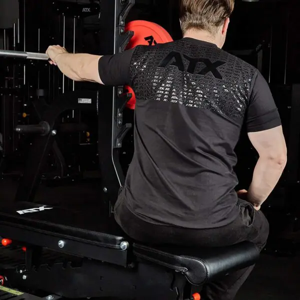 A person on a workout bench grips a barbell in a gym, wearing an ATX® Grip Shirt and getting ready for weightlifting, with gym equipment visible in the background.
