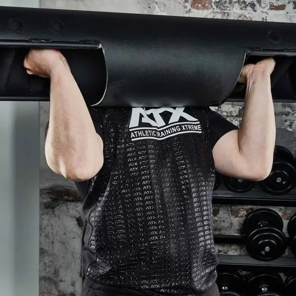 Someone wearing an ATX® Grip Shirt is lifting a heavy black weight bar. Only their arms and upper body are visible against a backdrop of gym equipment and a brick wall.