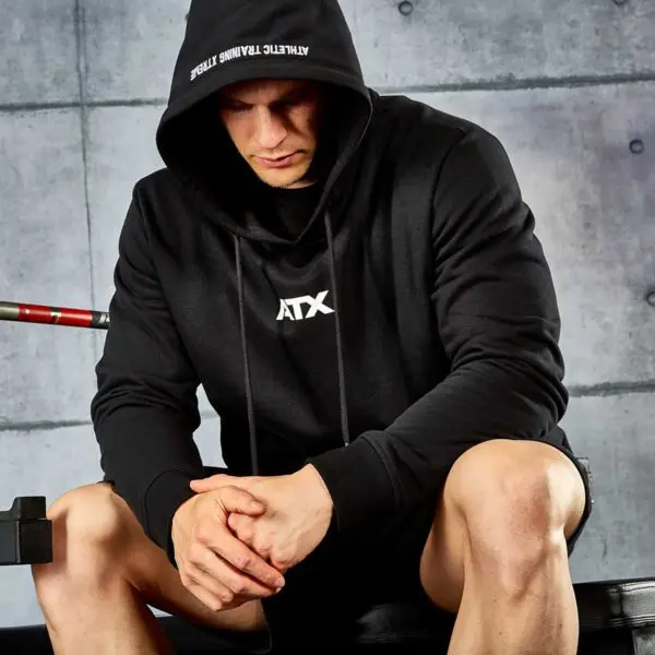Wearing an ATX® Black Hoodie, a person sits with clasped hands on a bench, gazing downward. A concrete wall forms the backdrop, with a barbell partly visible to the left.