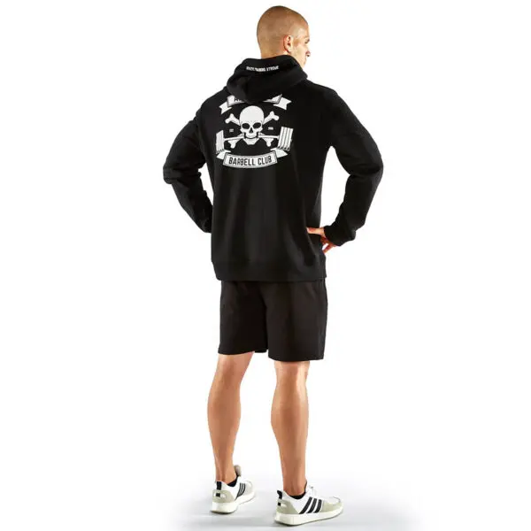 A man wearing an ATX® Black Hoodie with a skull and crossbones design and Barbell Club text on the back stands with his back to the camera, paired with black shorts and white sneakers featuring black stripes.