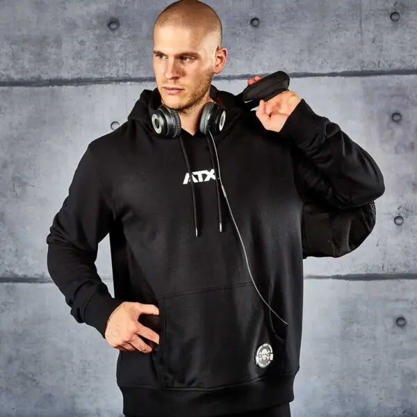 A man wearing an ATX® Black Hoodie stands against a concrete wall with headphones around his neck, holding a black device over his shoulder and looking off to the side.
