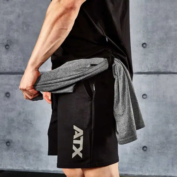 A person in a black shirt and ATX® Black Shorts, with ATX on the side, ties a gray shirt around their waist. A gray concrete wall with visible seams is in the background.