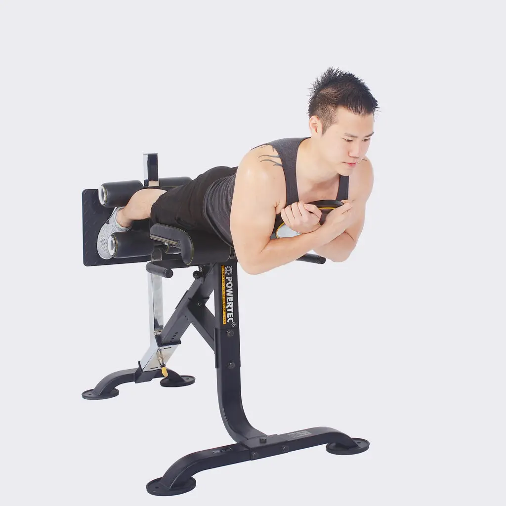 A person exercising on a Powertec Hyperextension bench, holding a small weight. The individual is positioned face down, with their torso parallel to the floor, engaged in a core strengthening exercise. They wear a tank top and shorts.