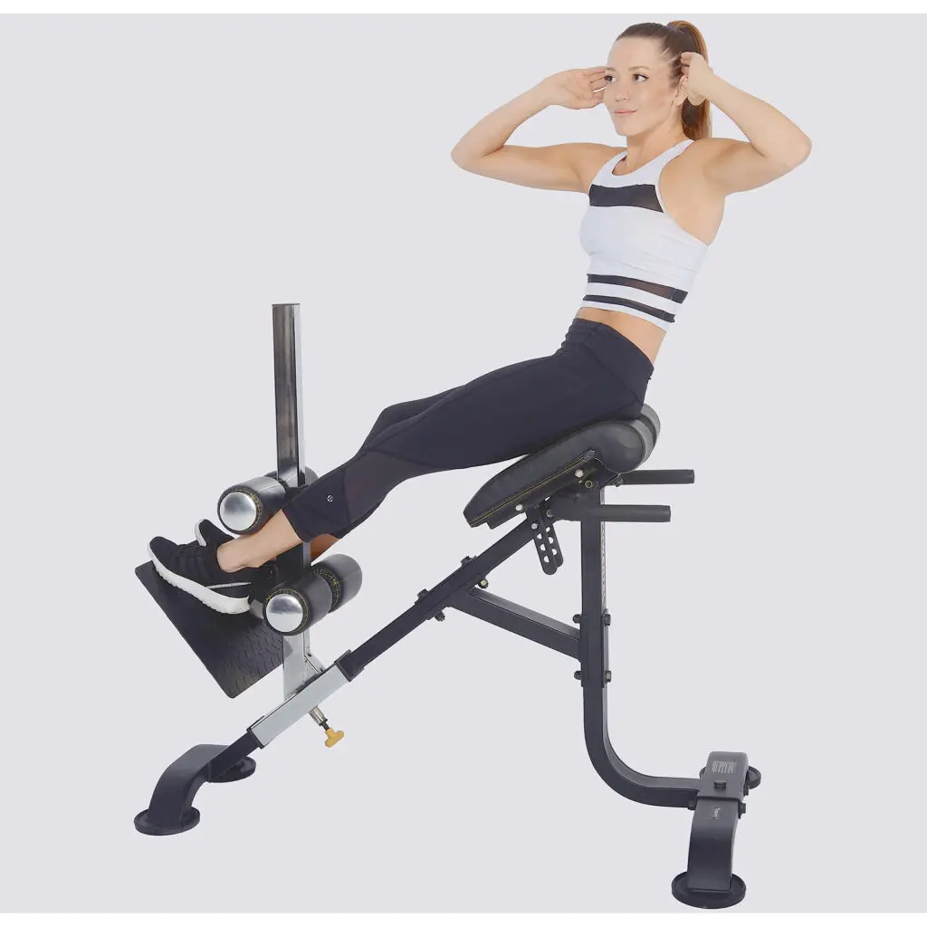 A woman in athletic wear is exercising on a slant board bench, performing sit-ups. Using the Powertec Hyperextension, her hands are by her head and feet secured under padded bars. The plain white background keeps the focus on her workout precision.