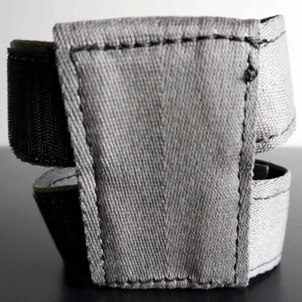A close-up of the Spud Inc 5R Barbell Shield shows a gray fabric strap with black stitching and a black elastic section, folded to display its texture. Its set on a smooth, dark surface.