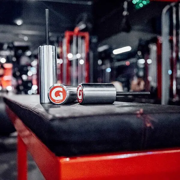 Close-up of two metallic GymPin 2 Original scent diffusers with red logo caps on a gym bench, set against gym equipment and blurred lights, creating a modern, energetic atmosphere.