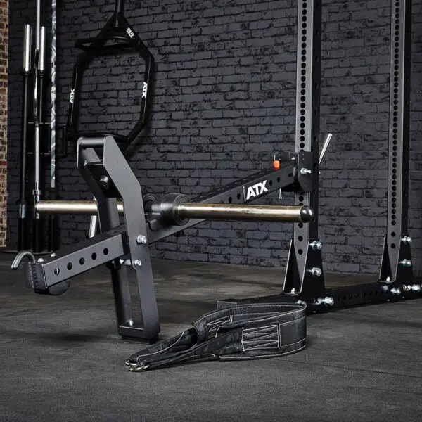 The ATX® Belt Squat Option features a weightlifting machine with a barbell, black padded straps, and adjustable metal arms. Designed for strength training against a dark brick wall, it includes secure bolts and a sturdy frame.