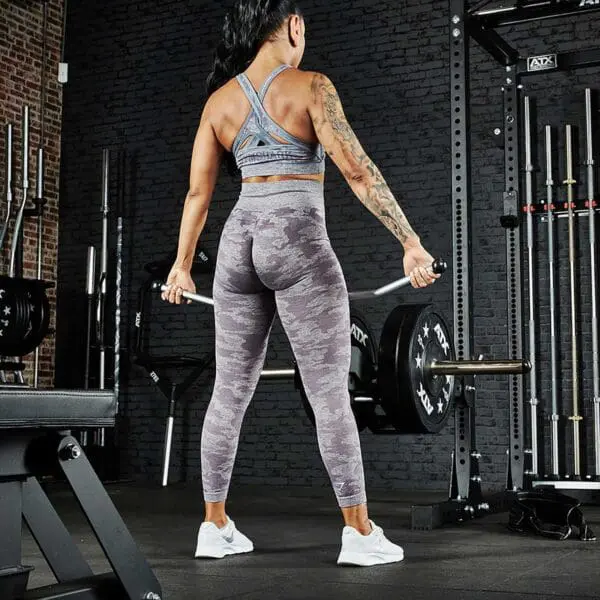 A person with long hair, wearing a sports bra and camouflage leggings, is lifting a barbell in a gym equipped with an ATX® Belt Squat Option. The space features brick walls and various workout gear like racks and benches.