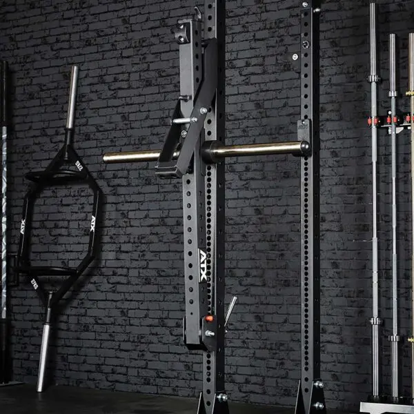 The gym setup showcases a black power rack and various barbells, including a trap bar and standard Olympic bars, against a dark brick wall. With the addition of the ATX® Belt Squat Option, it creates an organized and functional workout space.