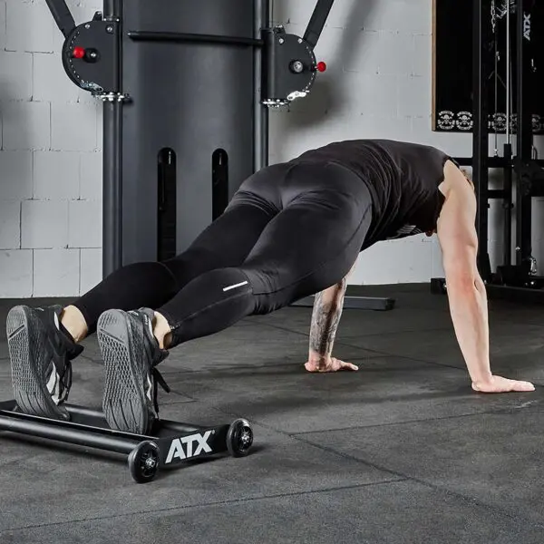 A person in a gym performs a decline push-up using a low bench, wearing black athletic clothing and shoes. The gym features various equipment, including the ATX® Glute Ham Roller visible in the background, with gray walls and rubber flooring.