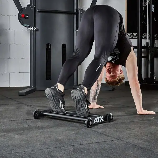 A person is performing a fitness exercise with the ATX® Glute Ham Roller in the gym. They are bent over with hands and feet on the floor, clad in black athletic clothing and shoes. Gym equipment, including a GHR machine, can be seen in the background.