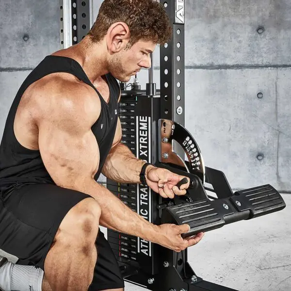 A man at a gym adjusts the ATX® Trinity Tower 135 kg. He wears a black tank top and shorts, concentrating on the machine. A concrete wall and a weights rack labeled Xtreme are visible in the background.