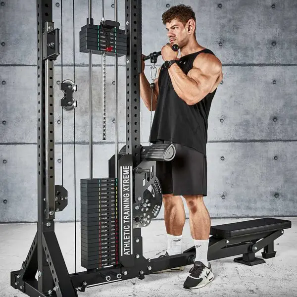 A muscular person in a black tank top and shorts performs a bicep curl on the ATX® Trinity Tower 135 kg cable machine in a gym, set against a concrete wall.