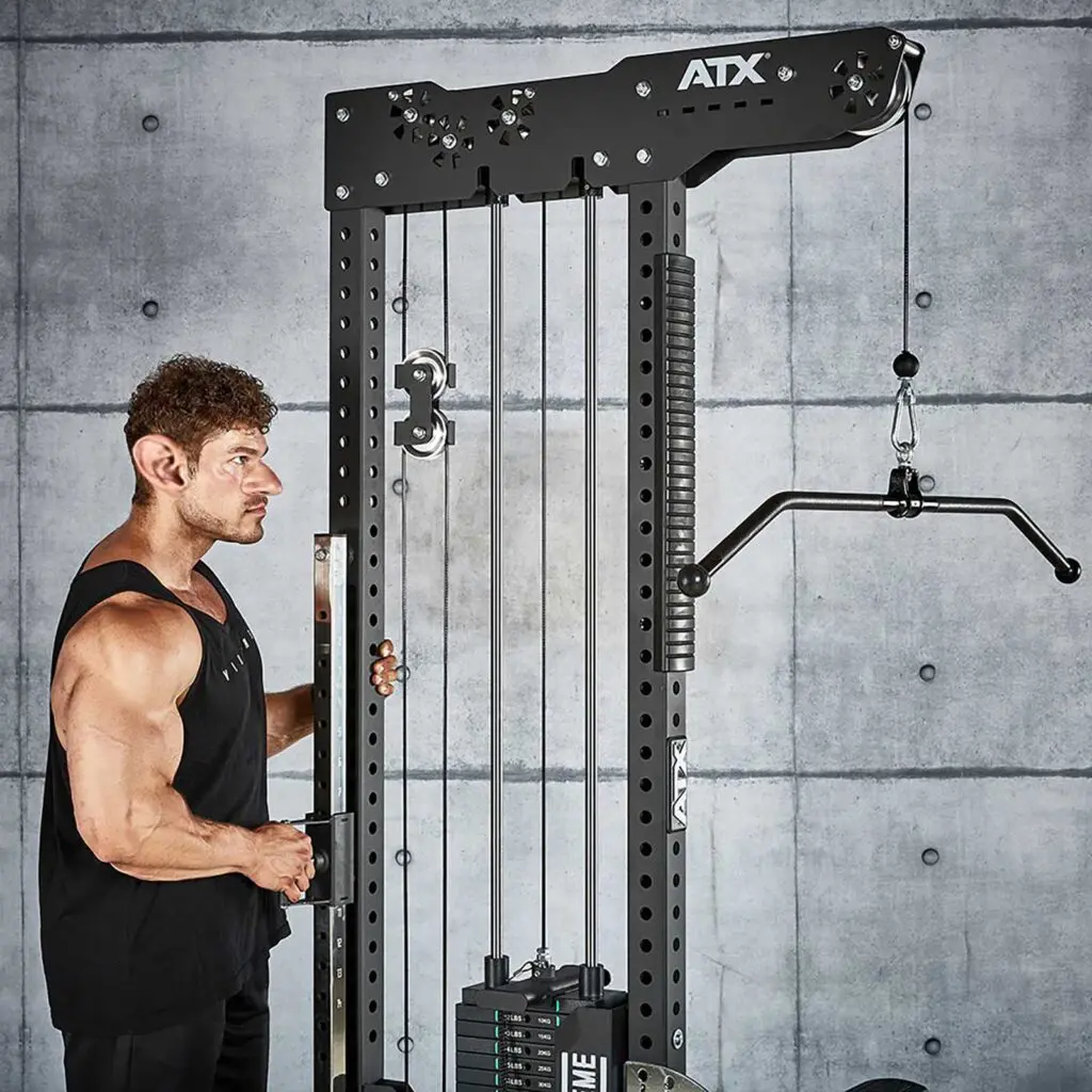 Wearing a black tank top, a person uses the ATX® Trinity Tower 135 kg, a tall black fitness machine with weights and pulley cables, against a concrete wall featuring a grid pattern.