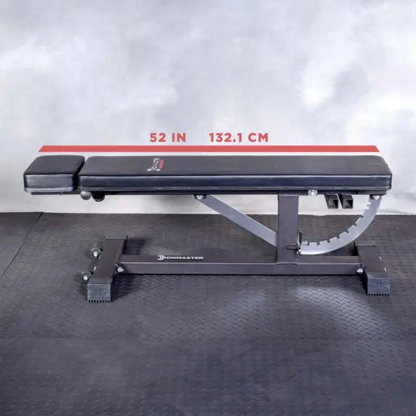 A black Ironmaster Head Extension SB adjustable gym bench sits on a black rubber floor against a gray wall, featuring a 52-inch (132.1 cm) measurement line on its frame.