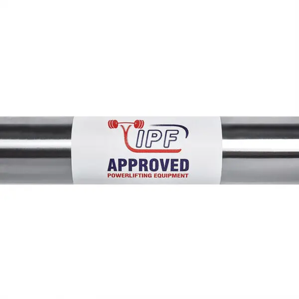 A close-up of a metallic bar with a white label displaying the IPF logo, featuring a red barbell graphic and the text APPROVED POWERLIFTING EQUIPMENT in bold red and blue letters on the ATX® Warrior Power Bar IPF.