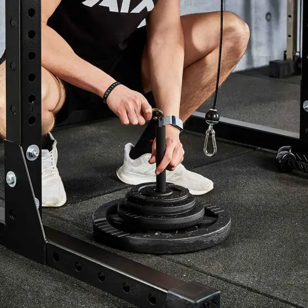 Next to the Cable Pulley System (High), a person in a black tank top, shorts, white sneakers, and a wristwatch adjusts weights on the loading pin. The gyms black mats cover the floor.