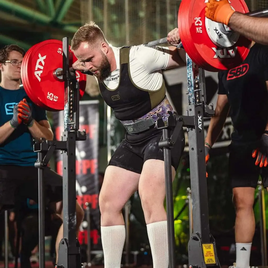 A man in a lifting suit prepares to squat with a loaded barbell on his shoulders during a gym competition, using the ATX® Combo Rack IPF Approved and assisted by gloved spotters on each side.