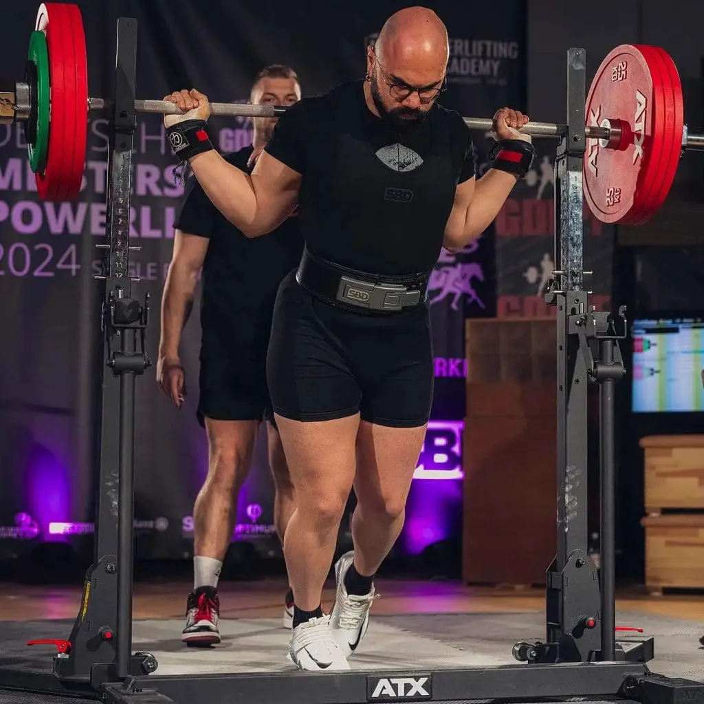 A powerlifter in a black outfit and belt prepares to squat a loaded barbell on the ATX® Combo Rack IPF Approved, with a spotter behind, in a gym setting.
