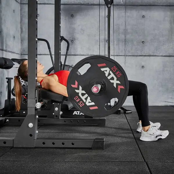 A long-haired person in a red top and black leggings skillfully performs a hip thrust exercise with a weighted barbell using the ATX® Hip Thruster Attachment. The gym features concrete walls and black flooring, and the weights are marked ATX 25 KG.