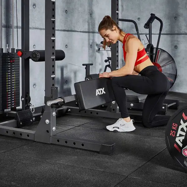 A woman in workout attire kneels to adjust an ATX® Hip Thruster Attachment on a gym machine. She wears a red sports bra, black leggings, and white sneakers. Other gym equipment is visible against the concrete wall in the background.