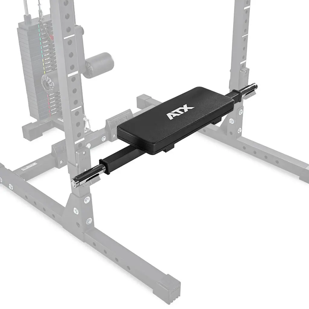 A gym setup includes a black bench pad with ATX printed on it, attached to an adjustable cable crossover machine. The ATX® Hip Thruster Attachment complements the sturdy metal frame and visible pulley system in the background.