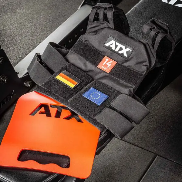 An ATX® Tactical Weight Vest in black, labeled 14 kg, adorned with German and EU flag patches, rests on gym equipment. Nearby on the floor is a red ATX-branded weight plate.