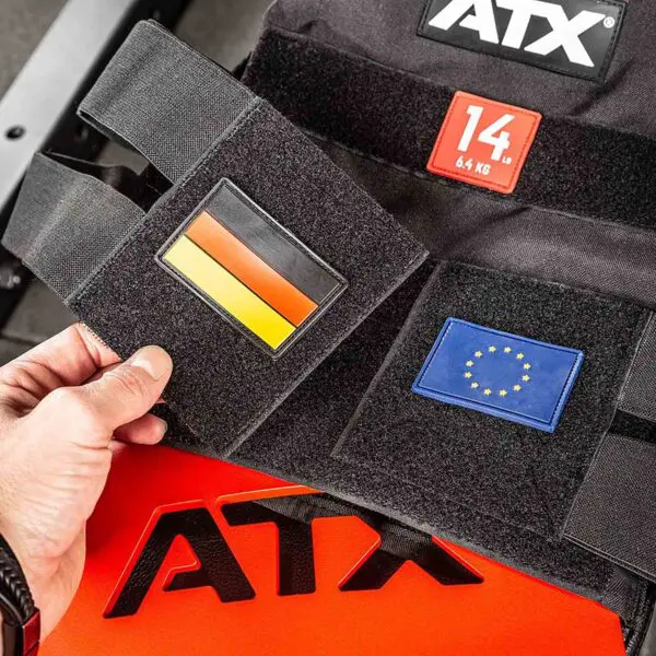 A hand holds a German flag patch on the ATX® Tactical Weight Vest, featuring another patch with the EU flag. A 14 kg weight label is visible. The vest rests on a red and black surface with ATX branding.