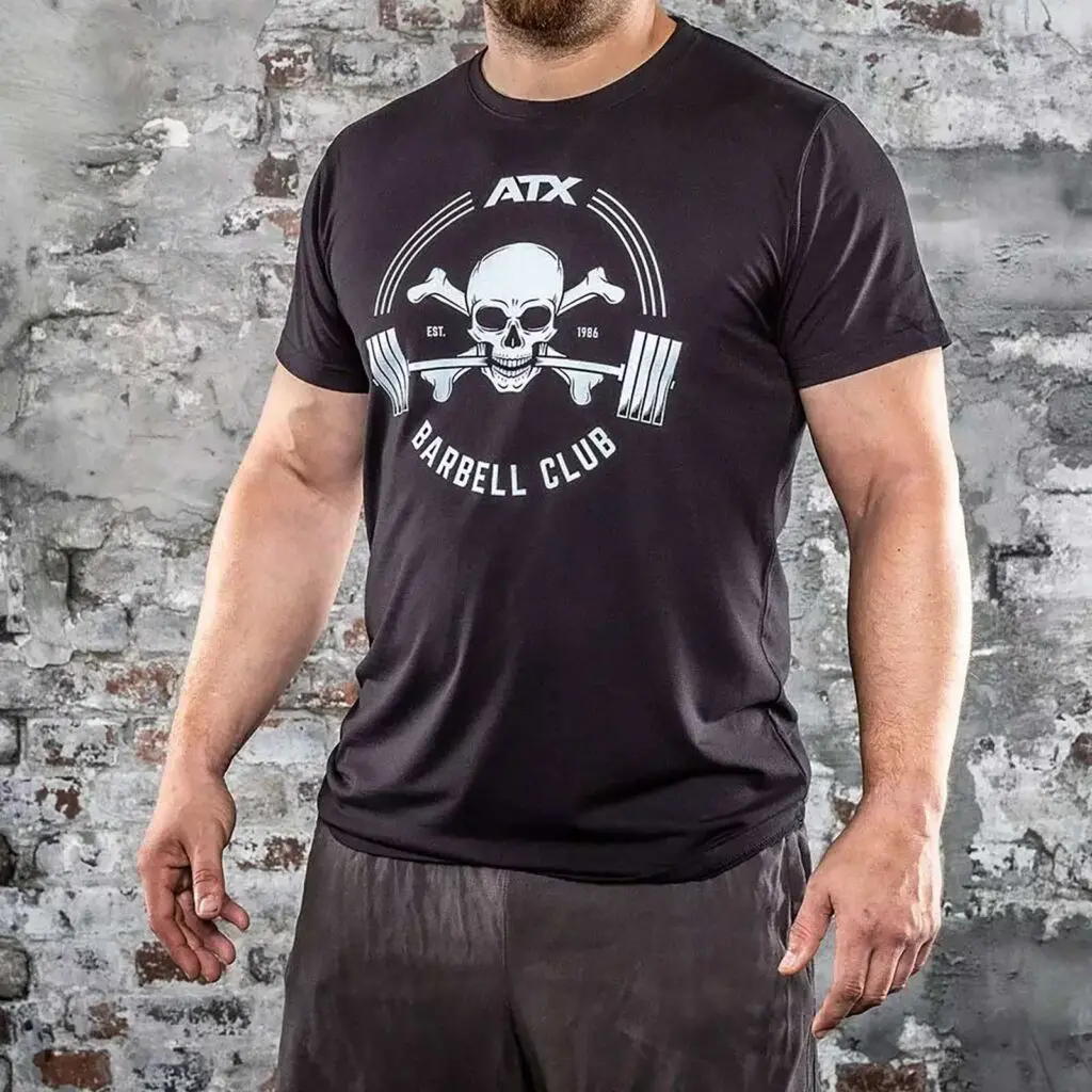 A person in black ATX® Barbell Club T-Shirt, featuring a skull and crossbones, stands in front of a weathered brick wall, paired with black shorts.