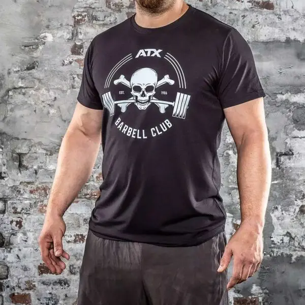A person in black ATX® Barbell Club T-Shirt, featuring a skull and crossbones, stands in front of a weathered brick wall, paired with black shorts.