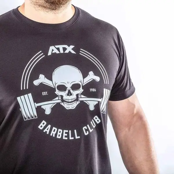 A person models the ATX® Barbell Club T-Shirt Black, showcasing a skull with crossed bones and a barbell in its mouth. The prints say ATX Barbell Club and Est. 1986. The shirt is displayed against a white background.