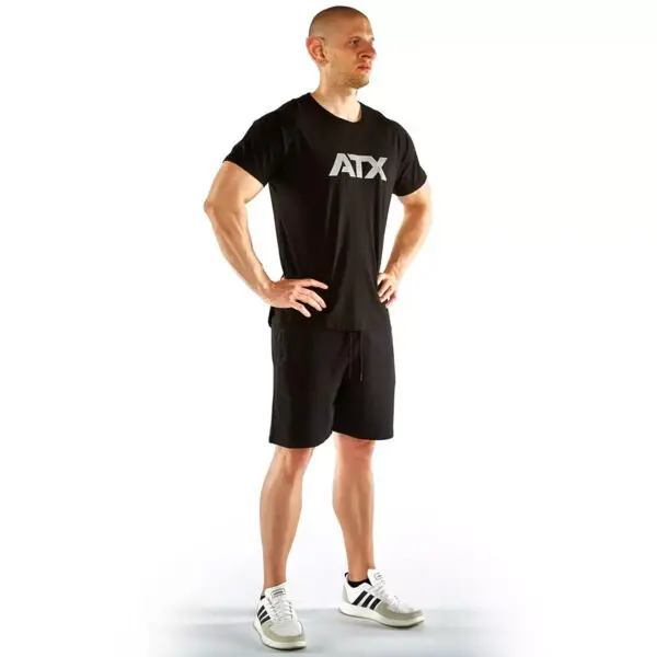 A person stands confidently with hands on hips, wearing a black ATX® Training Shirt, black shorts, and white Adidas sneakers against a plain white background.
