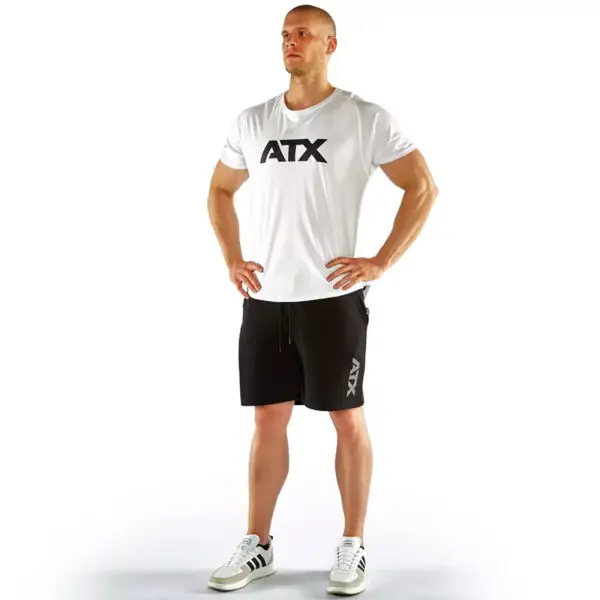 A person stands confidently with hands on hips, wearing the ATX® - T-Shirt White and black shorts with ATX branding. They sport white sneakers accented with black stripes, set against a plain white background.