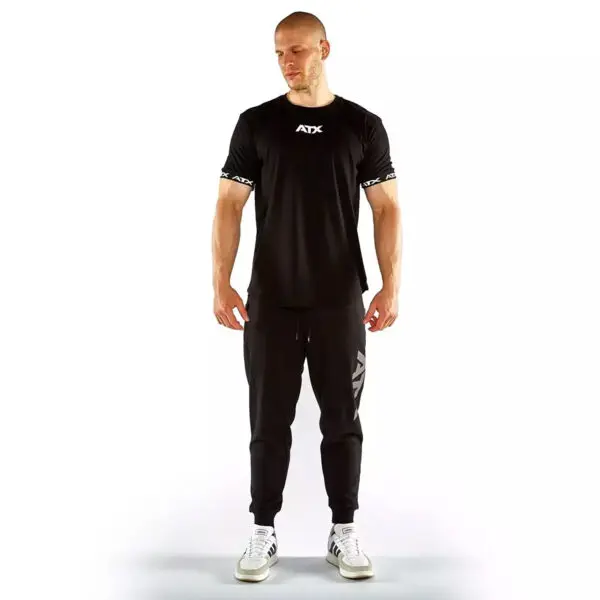 A man in a black ATX® Training Shirt BLACK, black joggers, and white sneakers stands against a plain white background. Hes bald with downcast eyes.