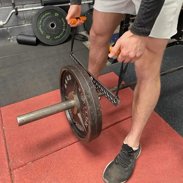 In a gym setting, someone in shorts and sneakers uses a hacksaw to cut a weightlifting bar with the T-BAR Row attachment Collar and weights in place, while another barbell with attached weights lies on the ground nearby.