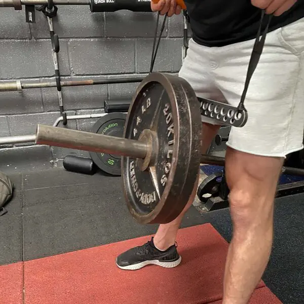 A person using a T-BAR Row attachment Collar with a weight plate, standing on a gym mat amid weightlifting bars and plates. They are wearing black shoes and white shorts.
